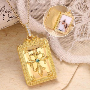 May include: Gold-colored rectangular locket necklace with a detailed floral design and a small blue gemstone. The locket opens to reveal a small photograph. The necklace is on a delicate gold chain.