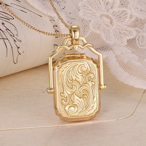 Könnte beinhalten: Eine goldfarbene rechteckige Anhängerhalskette mit einem verzierten floralen Design. Der Anhänger hängt an einer zarten Kette. Der Anhänger hat ein Scharnierdesign und das florale Muster ist auf der Vorderseite eingraviert.