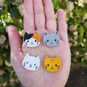 Può includere: Sono esposte quattro piccole statuette a forma di gatto. I gatti sono dipinti in bianco e nero, arancione e bianco, grigio, bianco e giallo con strisce arancioni. Ogni gatto ha una faccia semplice, in stile cartone animato. Le statuette sono tenute in una mano.
