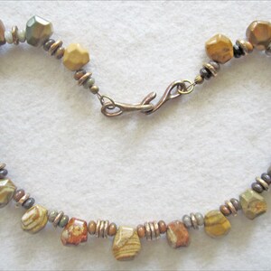 May include: A brown, green, and beige stone bead necklace with a copper clasp. The necklace is made of faceted beads and has a rustic look.