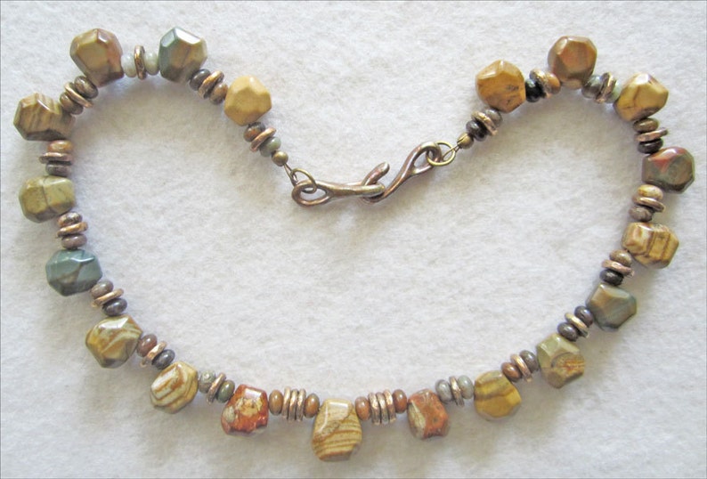 May include: A brown, green, and beige stone bead necklace with a copper clasp. The necklace is made of faceted beads and has a rustic look.