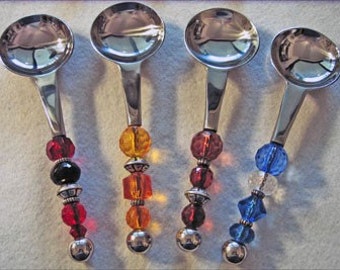 May include: Four decorative spoons with silver bowls and handles. Each handle features a unique design of colorful beads, including red, black, orange, and blue, separated by silver accents. The spoons are arranged on a white surface.