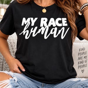 Puede incluir: Una camiseta negra de manga corta con el texto blanco MY RACE en una fuente sans-serif en negrita sobre la palabra human escrita en letra cursiva blanca. La camiseta se combina con unos vaqueros de mezclilla azul claro desgastados.