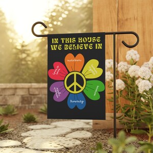 May include: A black garden flag with a rainbow peace sign in the center. The peace sign is surrounded by six rainbow hearts with the words "love is love", "inclusion", "human rights", "science", "social justice", and "diversity" written on them. The flag reads "In this house we believe in".