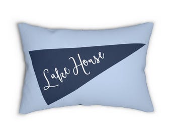 May include: A pale blue rectangular cushion featuring a navy blue pennant design. The pennant displays the words "Lake House" in white script. This cushion is a decorative item for the home.