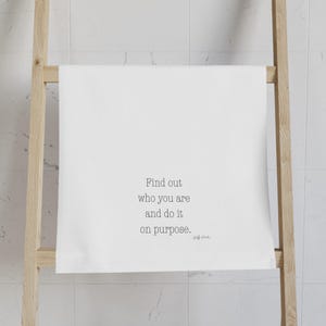 May include: White towel featuring the quote "Find out who you are and do it on purpose." in a minimalist font. The towel is hung on a wooden ladder-style rack against a white tiled wall.