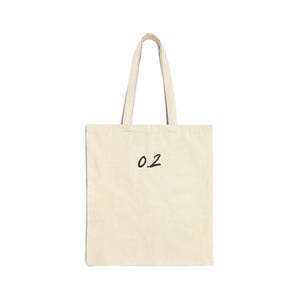 May include: Cream-colored tote bag with black text reading "0.2". The bag has two handles and is made of a sturdy fabric. The design is simple and minimalist, suitable for everyday use.
