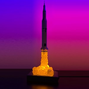 Peut inclure: Une fusée modèle blanche et grise avec une base à effet de flamme jaune. La fusée est sur une base noire et est éclairée d'une lumière jaune chaude.