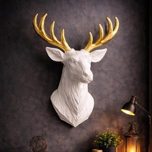 May include: A white deer head wall sculpture with gold-colored antlers. The deer head is mounted on a dark gray wall. The sculpture is a decorative piece for home decor.