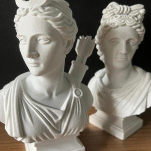 May include: Two white busts of classical figures. One bust features a crescent moon on its head and a quiver of arrows. The other bust has a detailed hairstyle. Both busts are on square pedestals and are set against a dark background.