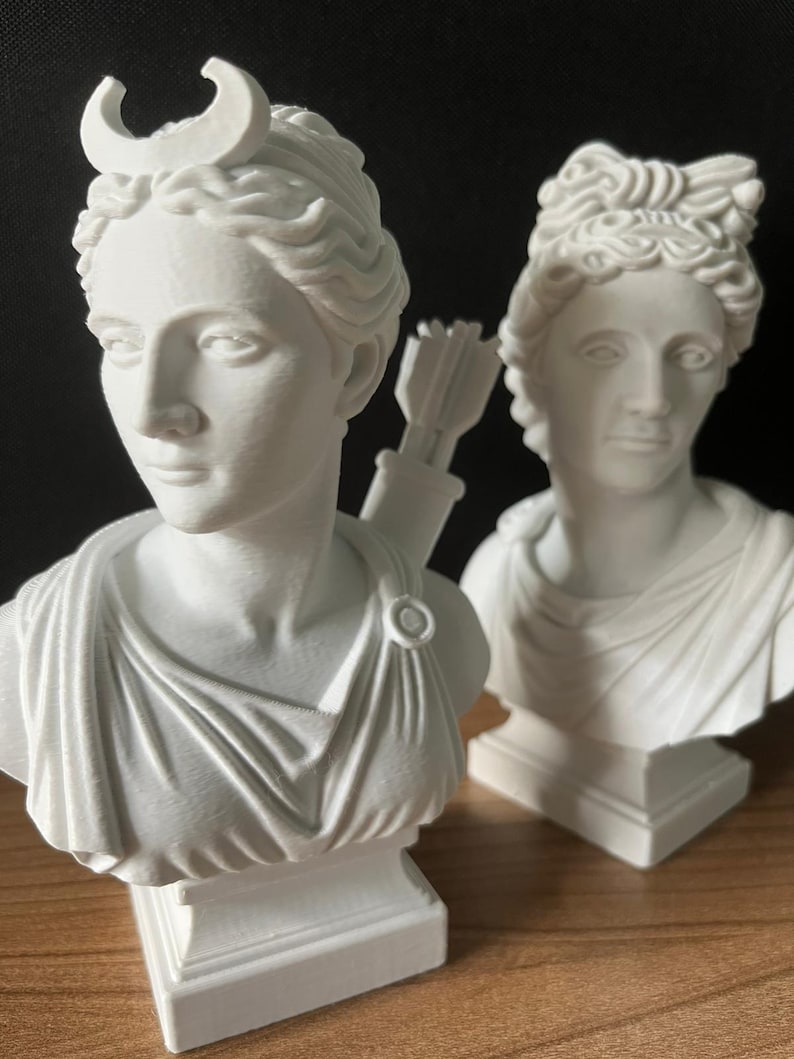May include: Two white busts of classical figures. One bust features a crescent moon on its head and a quiver of arrows. The other bust has a detailed hairstyle. Both busts are on square pedestals and are set against a dark background.