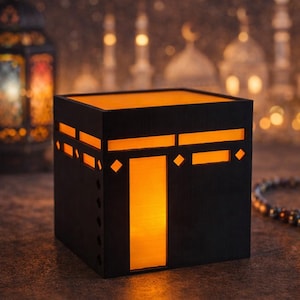 May include: A black, cube-shaped decorative lantern with an orange glow emanating from within. The lantern features cut-out geometric designs and is set against a blurred background with mosque silhouettes and hanging stars.