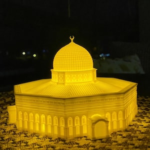 May include: A 3D printed model of a mosque with a golden dome and intricate details. The model is illuminated from within, casting a warm glow.