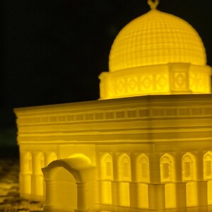 May include: A white 3D printed model of a mosque with a dome and intricate details. The model is illuminated from within, casting a warm yellow glow.