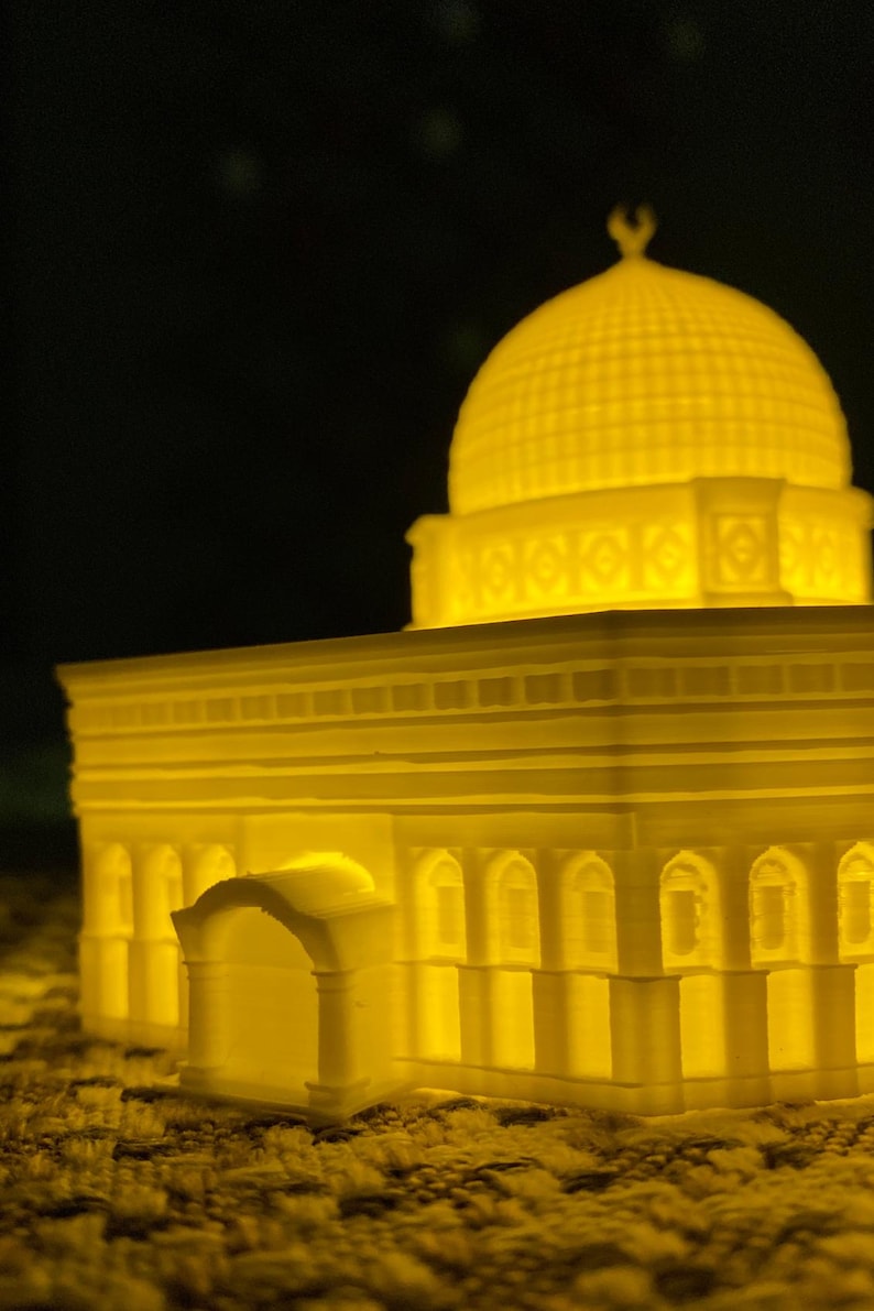 May include: A white 3D printed model of a mosque with a dome and intricate details. The model is illuminated from within, casting a warm yellow glow.
