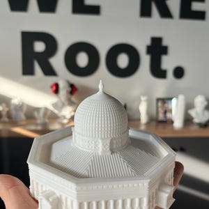May include: White 3D printed model of a building with a domed roof. The model is sitting on a hand.