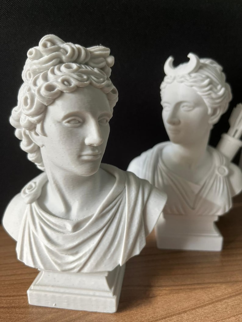 May include: Two white busts of classical figures. The bust on the left has curly hair and a draped garment. The bust on the right has a crescent moon in its hair and a quiver of arrows. Both are on square pedestals.