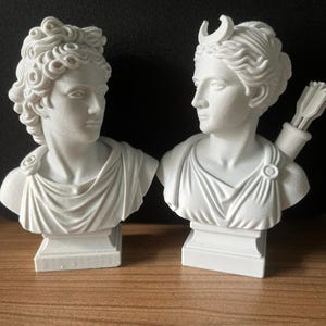 May include: Two white plaster busts of classical figures. One bust has curly hair and the other has a crescent moon on its head and a quiver of arrows. Both busts are on square pedestals and are facing each other.