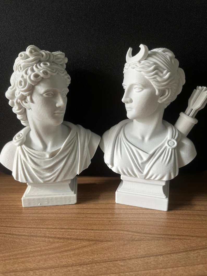 May include: Two white plaster busts of classical figures. One bust has curly hair and the other has a crescent moon on its head and a quiver of arrows. Both busts are on square pedestals and are facing each other.