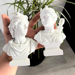 May include: Two white busts of classical figures. One bust has curly hair and a draped garment. The other bust has a bow and arrow and a draped garment. The busts are on a white base.