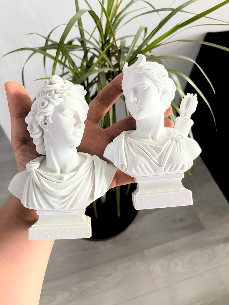 May include: Two white busts of classical figures. One bust has curly hair and a draped garment. The other bust has a bow and arrow and a draped garment. The busts are on a white base.
