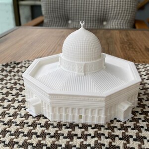 May include: White 3D printed model of a mosque with a domed roof and intricate details. The model is sitting on a patterned surface.
