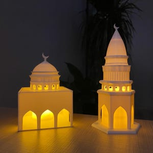 Peut inclure: Deux modèles de mosquée éclairés imprimés en 3D de couleur blanche. Le modèle de gauche a un dôme et des arches. Le modèle de droite est un minaret avec un sommet pointu.