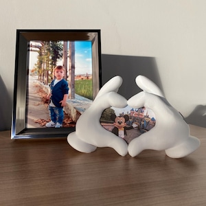 Peut inclure: Des mains en céramique blanche formant un cœur, tenant une photo de Mickey Mouse devant un château. La photo est dans un cadre blanc.