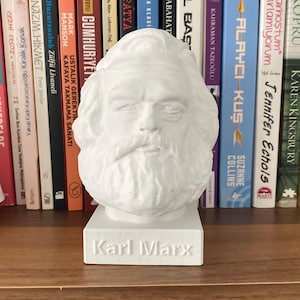 Może przedstawiać: Biała popiersie Karola Marksa na białej podstawie z napisem "Karl Marx" na przodzie. Popiersie stoi na drewnianej półce z rzędem książek z tyłu.