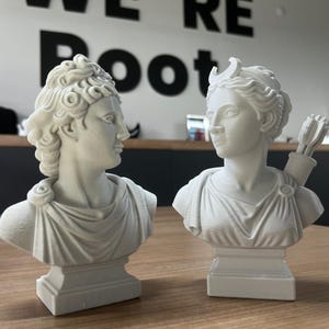 May include: Two white classical bust sculptures, one with a crescent moon and quiver of arrows. The busts are on a wooden table, with the words "WE'RE Root" in black on the wall behind them. The sculptures are detailed and have a classical aesthetic.