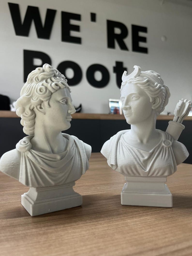 May include: Two white classical bust sculptures, one with a crescent moon and quiver of arrows. The busts are on a wooden table, with the words "WE'RE Root" in black on the wall behind them. The sculptures are detailed and have a classical aesthetic.