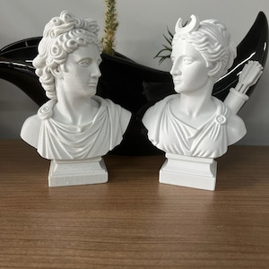 May include: Two white plaster busts of classical figures. One bust has curly hair, the other has a crescent moon on its head and a quiver of arrows. Both busts are on square pedestals.