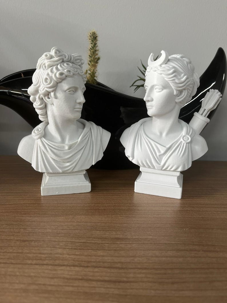 May include: Two white plaster busts of classical figures. One bust has curly hair, the other has a crescent moon on its head and a quiver of arrows. Both busts are on square pedestals.