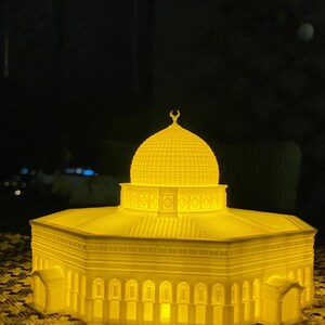 May include: A 3D printed model of a mosque with a golden yellow finish. The model is lit from within, creating a warm glow. The mosque has a domed roof and intricate details.
