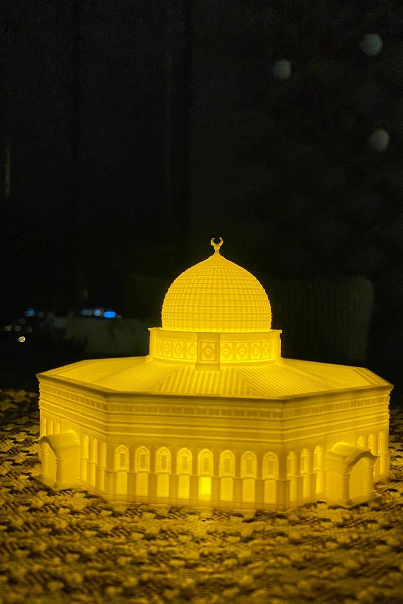 May include: A 3D printed model of a mosque with a golden yellow finish. The model is lit from within, creating a warm glow. The mosque has a domed roof and intricate details.
