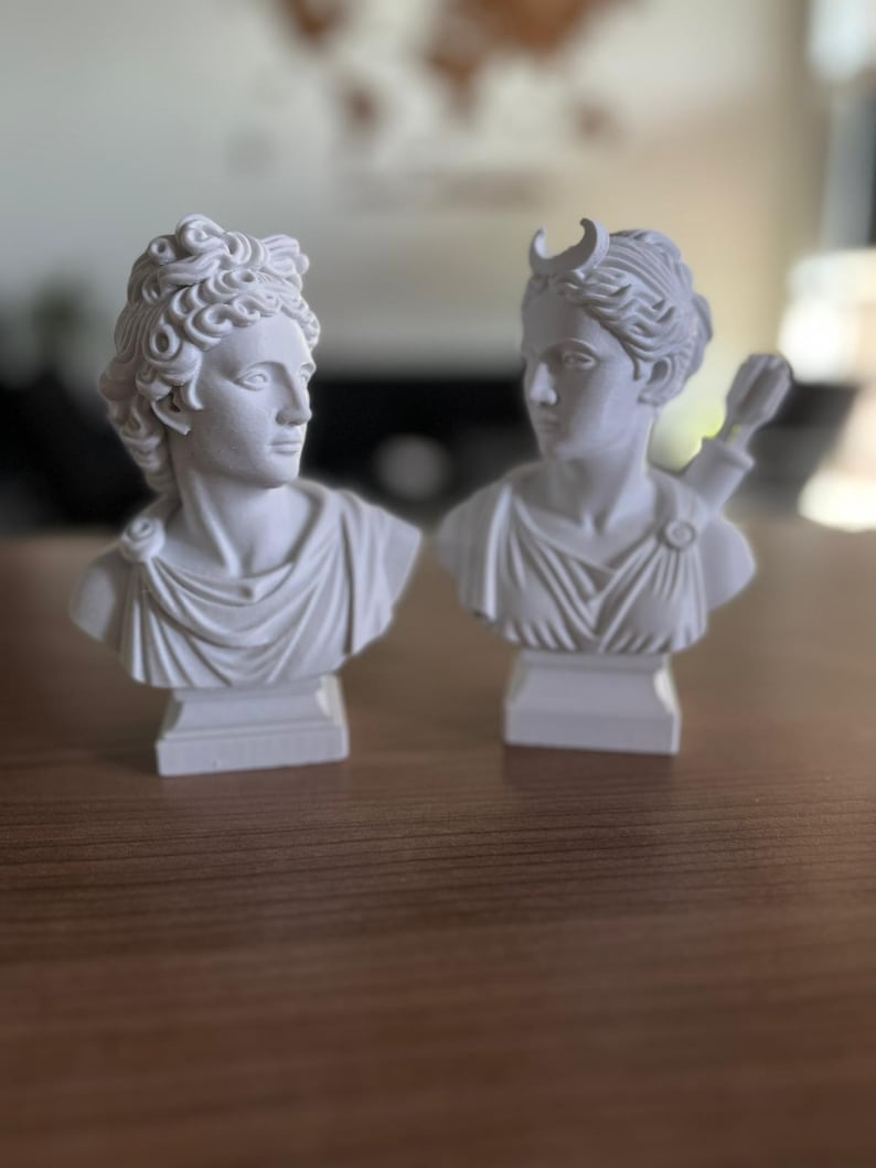 May include: Two white plaster busts of classical figures. One bust features a male figure with curly hair and a draped garment. The other bust depicts a female figure with a crescent moon on her head and a quiver of arrows.