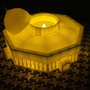 May include: A white 3D printed candle holder shaped like a dome with a removable top. The candle holder is lit and glowing yellow.
