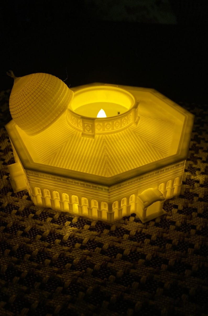 May include: A white 3D printed candle holder shaped like a dome with a removable top. The candle holder is lit and glowing yellow.