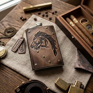 May include: A brown leather-wrapped metal flask featuring an embossed horse head design, secured with metal rivets. The flask includes two small leather tags, one labelled "hand made" and the other with an illegible brand name, attached by a string.