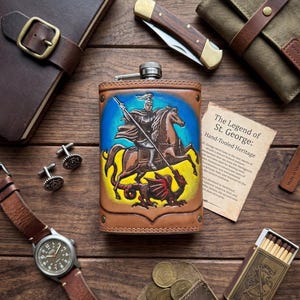 May include: A leather-wrapped metal flask featuring a hand-tooled and painted image of Saint George on horseback slaying a dragon. The flask is surrounded by a brown leather journal, a folding pocket knife, a watch, a matchbox, and a card.