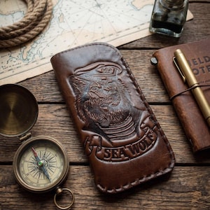 Puede incluir: Un estuche de cuero marrón oscuro con un diseño en relieve de un lobo con gorro de marinero fumando una pipa. Debajo del lobo, el texto SEA WOLF está grabado en una cinta. El estuche tiene costuras visibles a lo largo de los bordes.