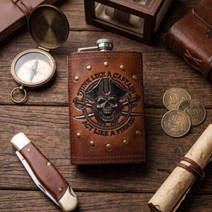 Op de afbeelding: Een met bruin leer beklede metalen heupfles met een reliëf van een piratenschedel met een kapiteinshoed, gekruiste zwaarden en de tekst "THINK LIKE A CAPTAIN ACT LIKE A PIRATE" omringd door kleine messingkleurige metalen klinknagels.