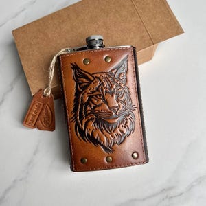 Op de afbeelding: Een met bruin leer omwikkelde metalen heupfles met een reliëf van een lynxkop op de voorzijde. De fles heeft een zilverkleurige schroefdop en is afgewerkt met vijf kleine messingkleurige metalen klinknagels. Een bruin label met hand made is bevestigd.