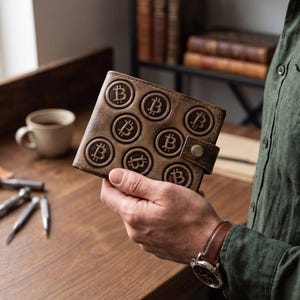 Può includere: Un portafoglio in pelle marrone con un motivo in rilievo di molteplici simboli Bitcoin circolari sulla superficie anteriore. Il portafoglio include una piccola chiusura a cinturino in pelle fissata con un singolo bottone a pressione in metallo color ottone.