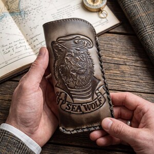 May include: A rectangular, tan-colored leather case featuring a detailed embossed design of a wolf wearing a sailor hat and smoking a pipe. Below the wolf, a banner displays the text SEA WOLF. The case has stitched edges and a smooth finish.