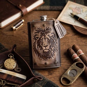 May include: A brown leather flask with a detailed embossed lion design. The flask has a silver cap and is accompanied by a small tag that reads "Hand Made". Other items include a pocket watch, cigars, and a cigar cutter.