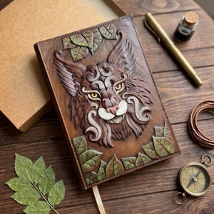 Puede incluir: Un diario de cuero rectangular que presenta un relieve tridimensional tallado a mano de una cara de lince con ojos amarillos. La cubierta está decorada con hojas verdes en relieve en las esquinas superiores e inferiores, en tonos marrones y beige.