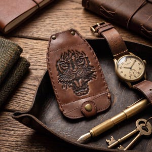 Puede incluir: Un estuche para llaves de cuero marrón con un diseño de cabeza de tigre en relieve, asegurado con broches de metal color latón. El artículo se muestra junto a un reloj de pulsera estilo vintage con correa de cuero marrón, una pluma de latón y dos llaves.