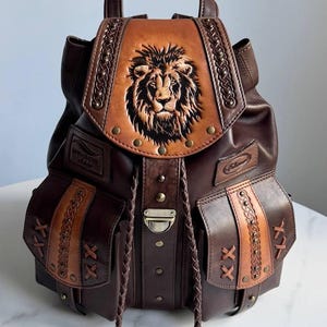 Op de afbeelding: Bruine leren rugzak met een gedetailleerd leeuwenkopontwerp op de voorklep. De rugzak heeft meerdere zakken, decoratieve stiksels en gevlochten trekkoorden. Hij heeft een handvat aan de bovenkant en verstelbare bandjes.