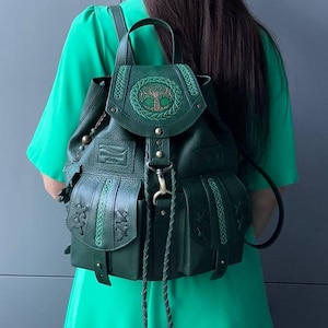 Puede incluir: Una mochila de cuero verde oscuro con un diseño circular de árbol en relieve en la solapa superior, dos bolsillos delanteros con detalles de cuero trenzado, un asa superior y largos cordones trenzados que cuelgan del cierre metálico central.